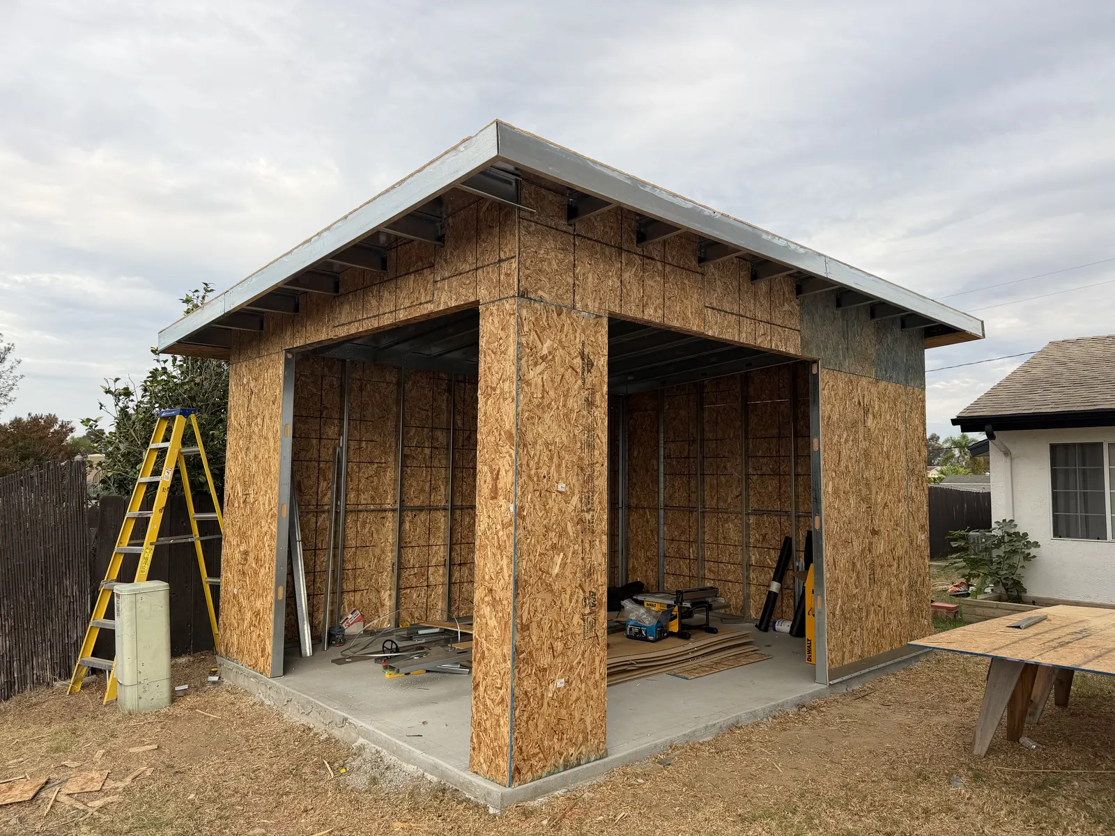 Front corner of a detached structure under construction, with two wide openings, visible metal framing, wall sheathing, and roof overhangs