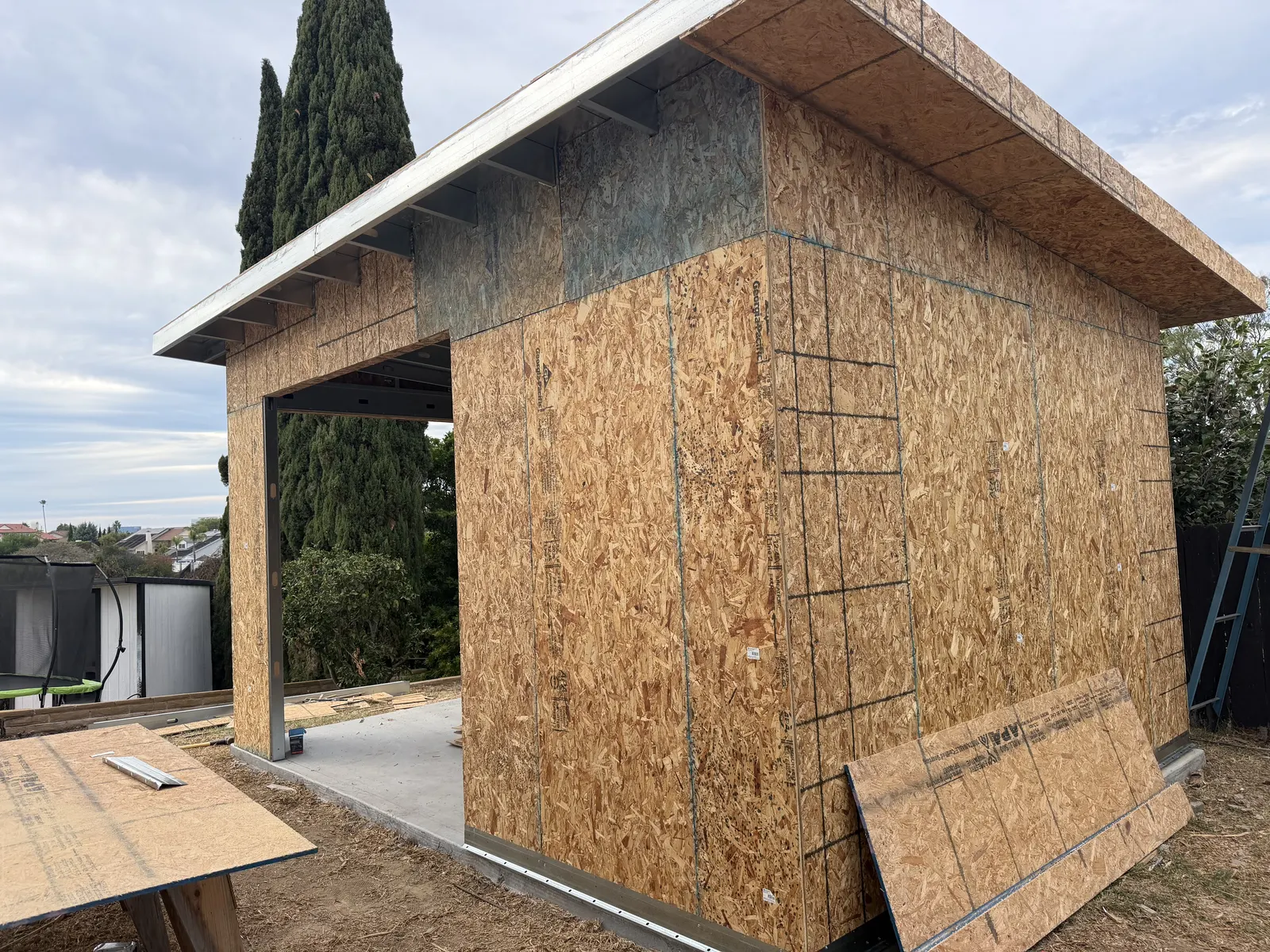 Side corner of the unfinished structure with wall sheathing, one open bay, and a projecting roof overhang