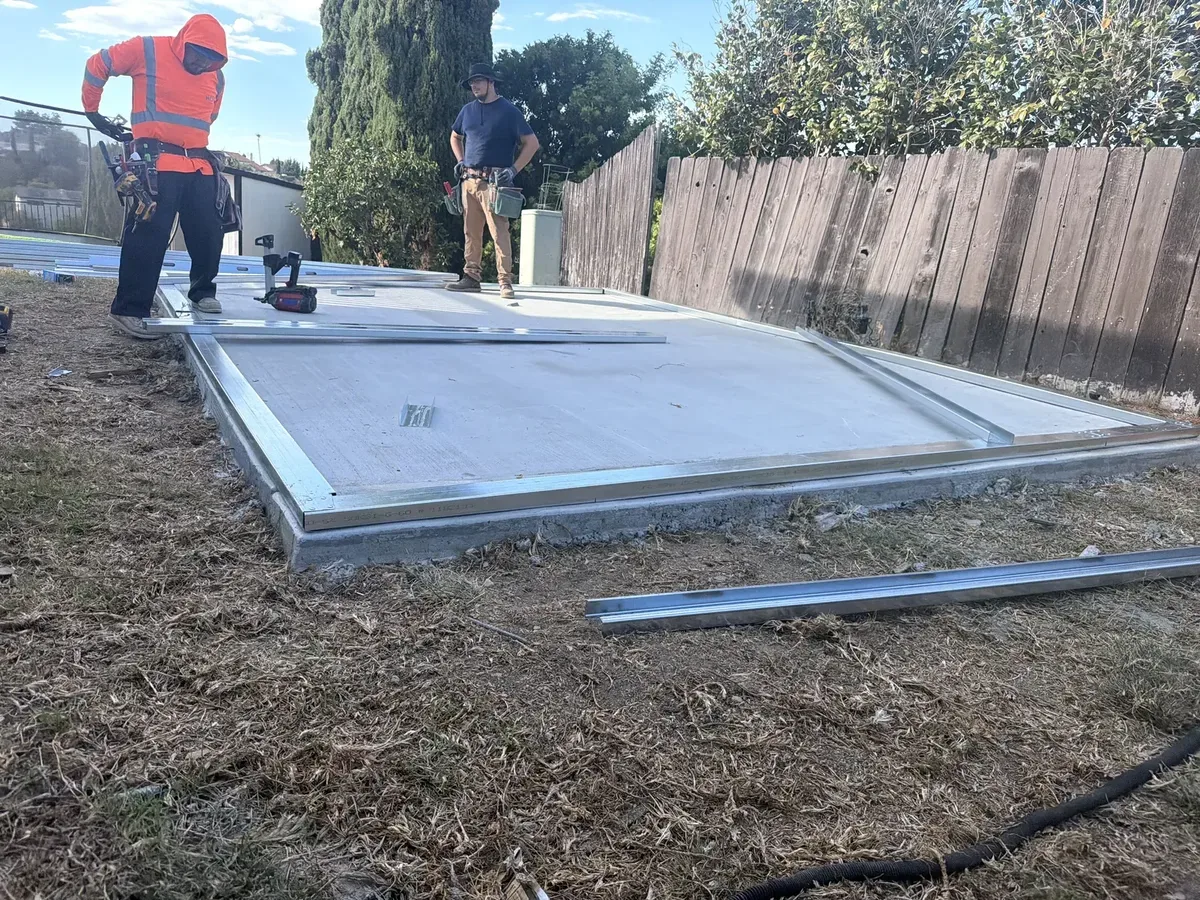 Two workers positioning metal framing pieces laid flat on a concrete slab at the unfinished structure